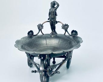 May include: An antique silver-toned metal serving dish in the shape of a wheelbarrow. The dish has a scalloped edge and is decorated with floral accents and a small statue of a woman. The wheelbarrow has three wheels.