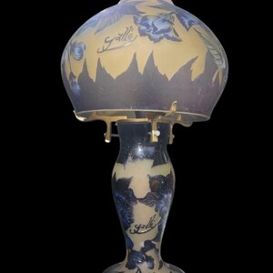 May include: A vintage table lamp with a floral design. The lamp features a glass shade with blue flowers and a gold background. The base is dark blue with a matching floral pattern. The lamp has the word "Galle" written on it.