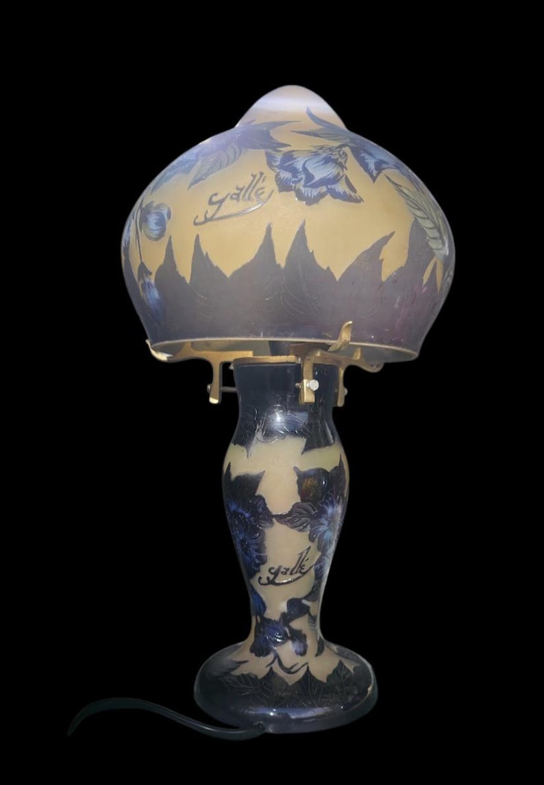 May include: A vintage table lamp with a floral design. The lamp features a glass shade with blue flowers and a gold background. The base is dark blue with a matching floral pattern. The lamp has the word "Galle" written on it.