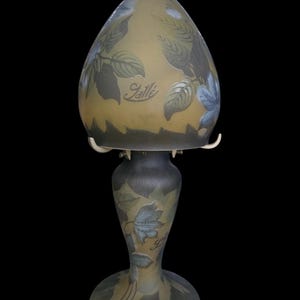 May include: An antique art glass lamp with a teardrop-shaped shade and a pedestal base. The lamp features a leaf design in shades of blue, green, and brown. The word "Galle" is inscribed on the shade and base.