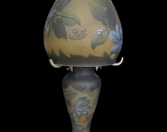 May include: A vintage art glass lamp with a teardrop-shaped shade and a pedestal base. The lamp features a leaf design in shades of blue, green, and brown. The word "Galle" is inscribed on the shade and base.