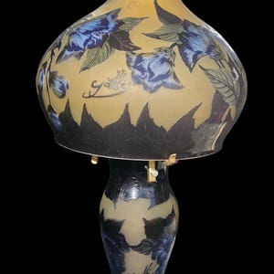 May include: A decorative table lamp with a floral design. The lamp features a yellow and blue glass shade with blue flowers and green leaves. The base is black with blue floral accents. The word "Galle" is written on the shade and base.