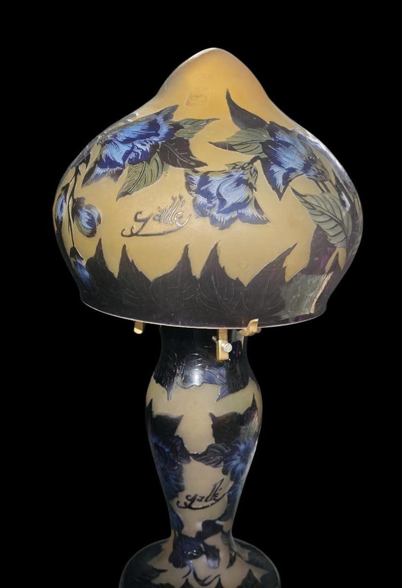 May include: A decorative table lamp with a floral design. The lamp features a yellow and blue glass shade with blue flowers and green leaves. The base is black with blue floral accents. The word "Galle" is written on the shade and base.