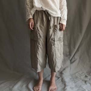 Può includere: Un paio di pantaloni larghi in lino color beige chiaro con vita a coulisse e grandi tasche a toppa. I pantaloni hanno un aspetto usurato e vintage con cuciture visibili e bordi sfilacciati.