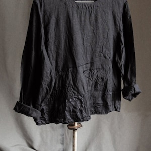 May include: A black linen shirt with a textured surface and a round neckline hangs on a metal hanger. The shirt has long sleeves and a relaxed fit.