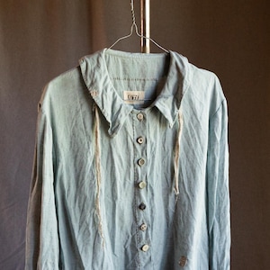 Op de afbeelding: Een lichtblauw linnen shirt met een kraag, lange mouwen en een knoopsluiting aan de voorkant. Het shirt heeft een rafelige zoom en een klein geborduurd ontwerp aan de voorkant. Het shirt hangt aan een hanger.