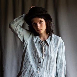 Könnte beinhalten: Eine Person trägt ein hellblaues, leicht abgenutztes, langärmliges Hemd mit Knopfleiste und einen schwarzen Hut. Das Hemd hat einen ausgefransten Saum und eine weiße Schnur, die um den Kragen gebunden ist.