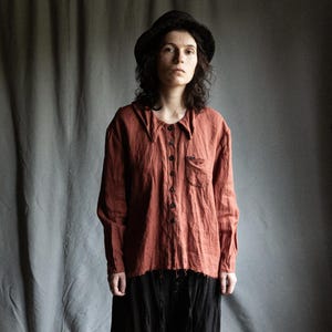 Könnte beinhalten: Eine Person trägt ein rostfarbenes Leinenhemd mit einem schwarzen Hut und einer schwarzen Hose.