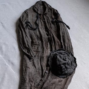 May include: A long, dark gray linen coat with a matching hat. The coat has a button-down front, two pockets, and long sleeves. The hat is a dark gray, textured fabric. The coat and hat appear to be handmade and have a rustic, vintage aesthetic.