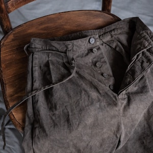Pode incluir: Uma calça cinza escura com cintura de cordão e fecho de botão. A calça está dobrada e colocada em uma cadeira de madeira.