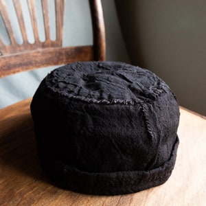 Puede incluir: Un sombrero negro de tela con un ala ancha y una superficie texturizada. El sombrero está sobre una superficie de madera.