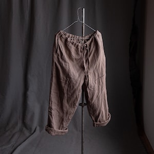 Peut inclure: Pantalon en lin marron avec une taille élastique et des ourlets retroussés, suspendu à un cintre métallique. Le pantalon décontracté présente des coutures visibles. Le fond est un tissu gris foncé.