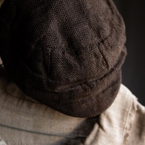 May include: A dark brown, woven fabric cap with a rounded crown and a flat brim. The cap is made of a thick, textured material.