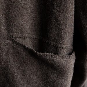 May include: Close-up of a dark brown, textured fabric with a visible pocket. The pocket is outlined with a contrasting stitch, adding a subtle detail to the garment. The fabric appears to be a soft, possibly woolen material.