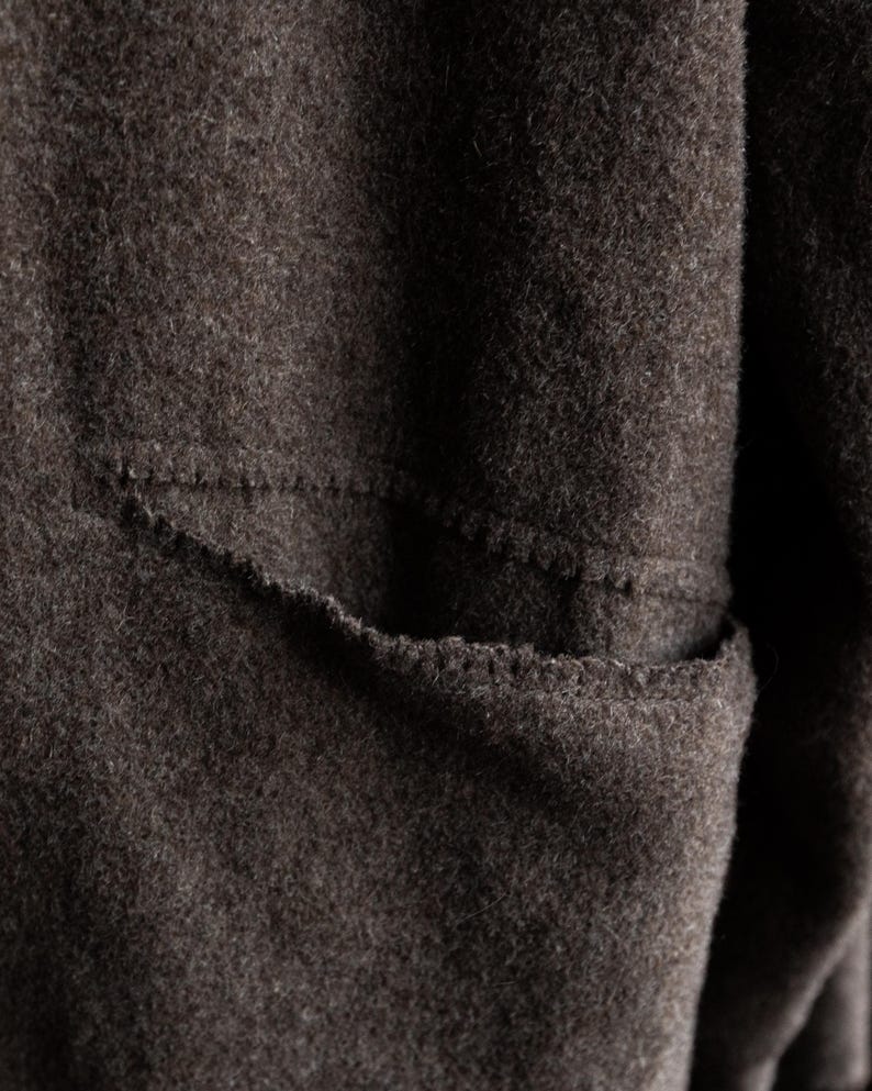 May include: Close-up of a dark brown, textured fabric with a visible pocket. The pocket is outlined with a contrasting stitch, adding a subtle detail to the garment. The fabric appears to be a soft, possibly woolen material.