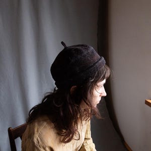 May include: A person wearing a brown felt beret with a pointed top. The person is wearing a light brown linen shirt and has long brown hair.