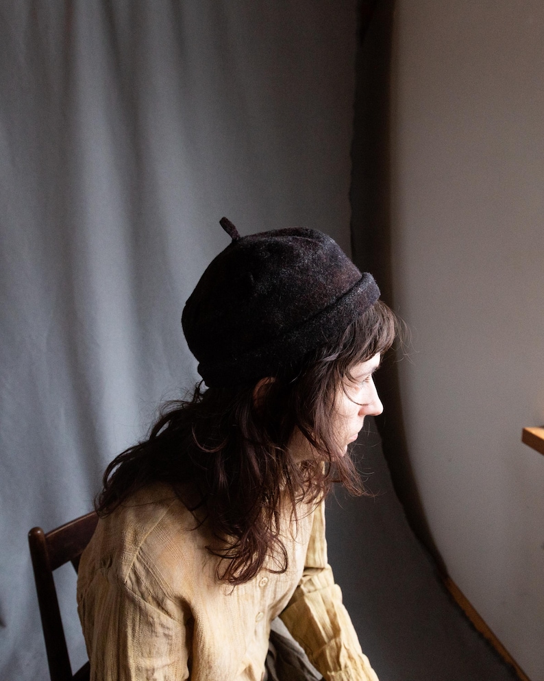 May include: A person wearing a brown felt beret with a pointed top. The person is wearing a light brown linen shirt and has long brown hair.
