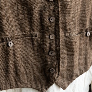 Può includere: Un gilet di lino marrone con una fila di sei bottoni marroni sul davanti. Il gilet ha due tasche sul davanti e un fondo a punta.