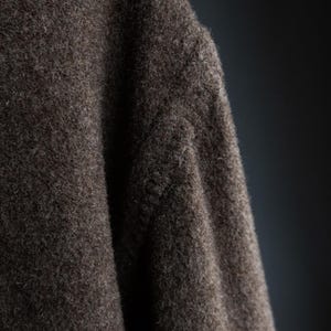 May include: Close-up of a section of a dark brown, textured fabric. The material appears to be a thick wool or similar textile, with a slightly uneven surface and visible fibers. The lighting creates shadows.