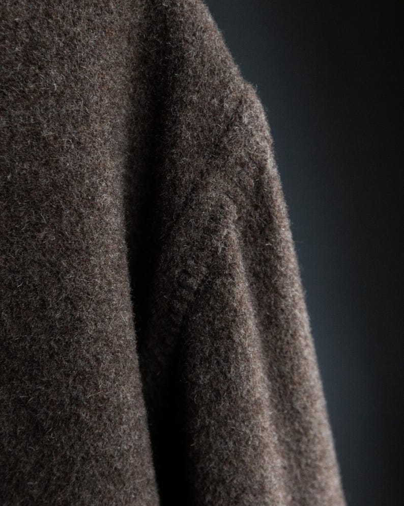 May include: Close-up of a section of a dark brown, textured fabric. The material appears to be a thick wool or similar textile, with a slightly uneven surface and visible fibers. The lighting creates shadows.