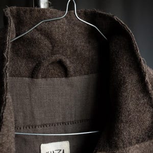 May include: Close-up of a brown wool coat collar hanging on a wire hanger. The coat has a textured appearance and a label that reads "KINZA". The coat is a neutral color and appears to be made of a thick material.