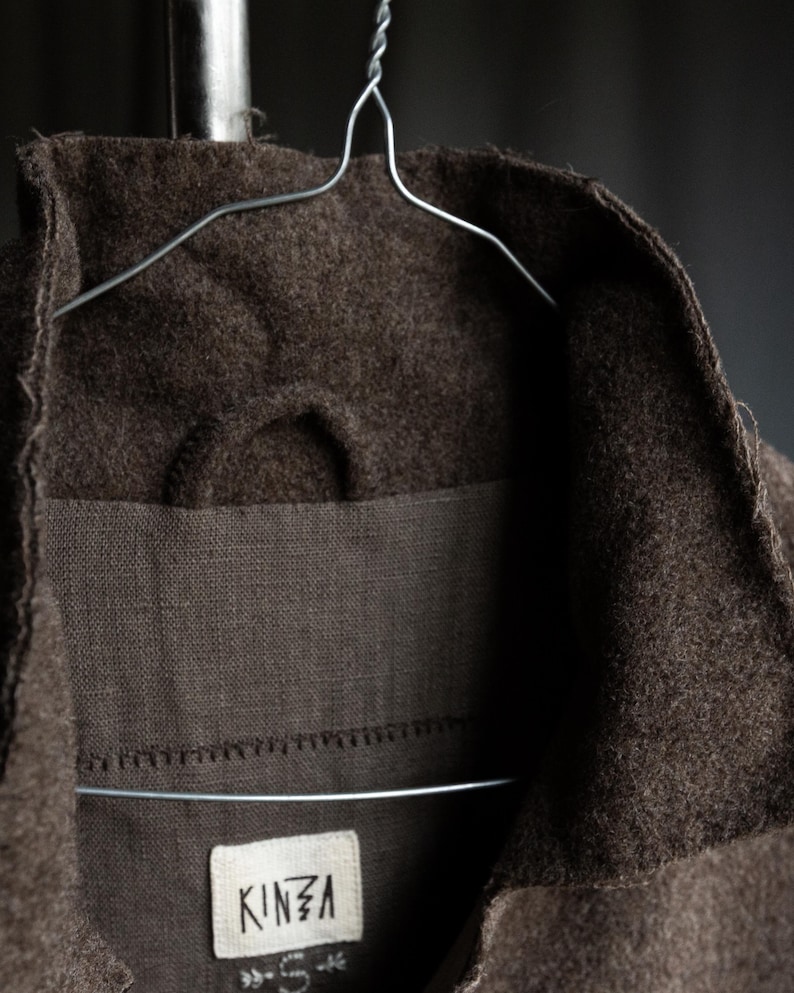 May include: Close-up of a brown wool coat collar hanging on a wire hanger. The coat has a textured appearance and a label that reads "KINZA". The coat is a neutral color and appears to be made of a thick material.