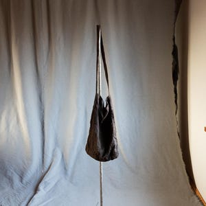 Può includere: Una borsa tote in tessuto grigio scuro appesa a un supporto metallico su uno sfondo bianco.