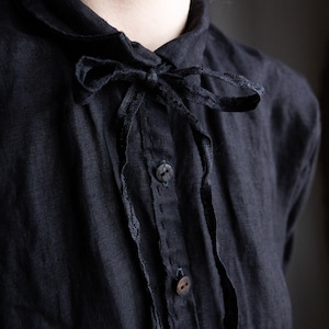 以下が含まれることがあります： 襟元にフリルが付いた黒いリネンシャツ。黒いリボンがボウタイになっていて、首元を飾っています。シャツには黒いボタンが2つ付いています。