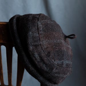 May include: A brown and grey wool beret with a small loop on the side. The beret is hanging on a wooden chair.