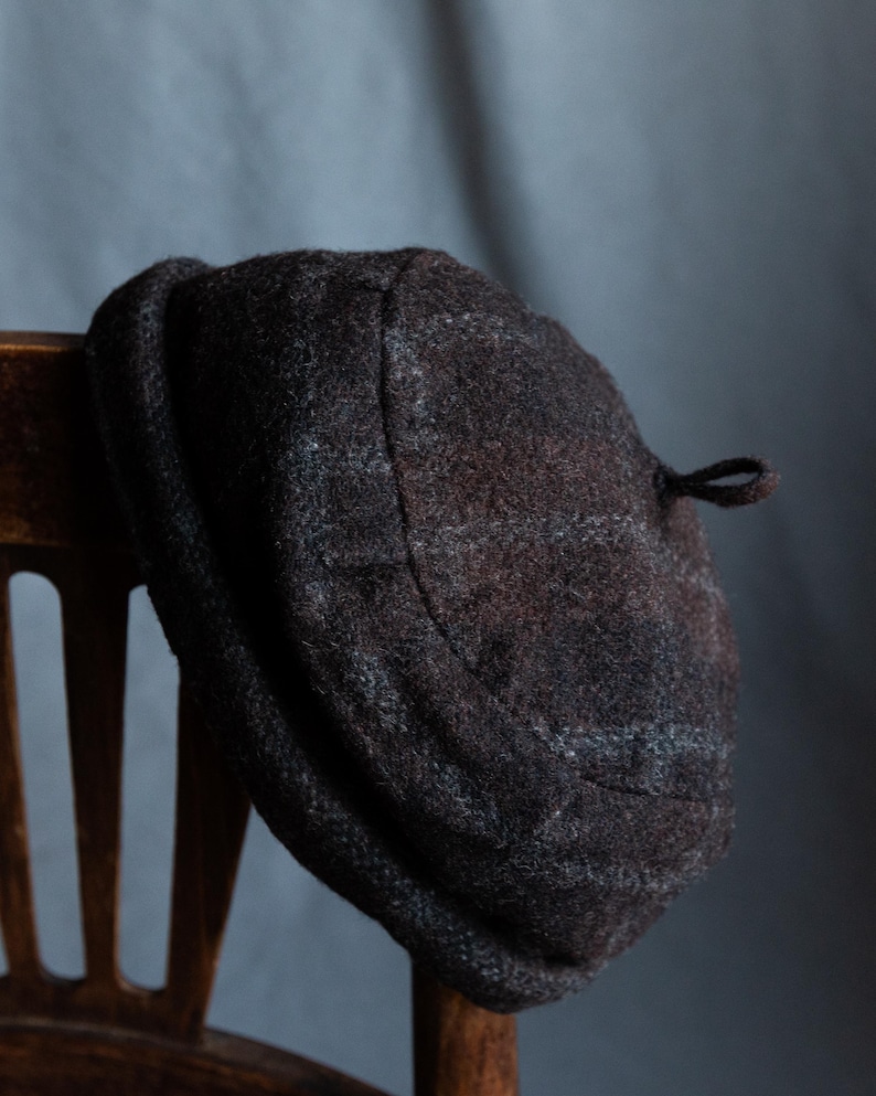 May include: A brown and grey wool beret with a small loop on the side. The beret is hanging on a wooden chair.
