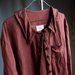 Könnte beinhalten: Ein rostfarbenes Leinenhemd mit einem großen Kragen und Knopfleiste. Das Hemd hat eine kleine Tasche auf der linken Seite und ein Etikett mit der Aufschrift "KUNZI".