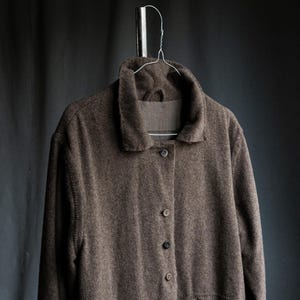 May include: A brown, button-down coat with a collar, hanging on a silver hanger. The coat has a textured appearance, with visible pockets and buttons down the front. The coat is made of a soft material.
