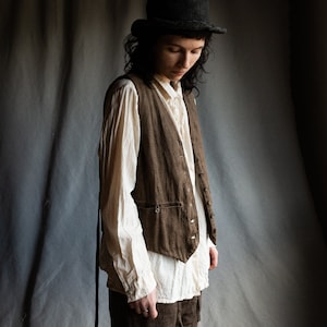 以下が含まれることがあります： 白いボタンダウンシャツと茶色のズボンを着た人が、茶色のリネンベストを着ています。その人は黒い帽子をかぶっています。