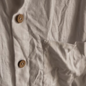 Könnte beinhalten: Nahaufnahme eines weißen Leinenhemdes mit zwei Holzknöpfen. Der Stoff ist zerknittert und hat eine natürliche Textur.