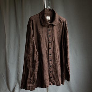 May include: A brown button-down shirt with a rounded collar, hanging on a metal hanger. The shirt has long sleeves and a series of dark buttons down the front. The fabric appears to be linen or a similar lightweight material.