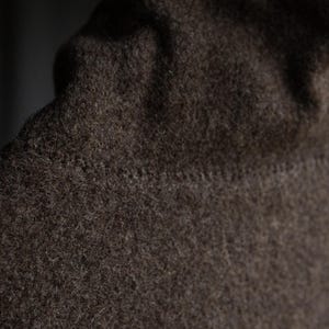 May include: Close-up of a dark brown, textured fabric, possibly wool or a similar material. The fabric has a slightly uneven surface and a visible seam. The image focuses on the texture and color of the material.
