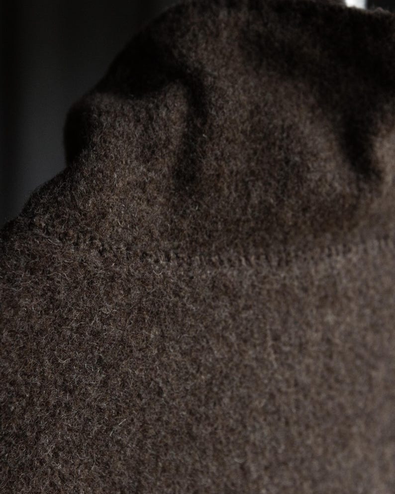 May include: Close-up of a dark brown, textured fabric, possibly wool or a similar material. The fabric has a slightly uneven surface and a visible seam. The image focuses on the texture and color of the material.