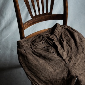 May include: Brown linen pants with a button fly and a textured weave. The pants are draped over a wooden chair.