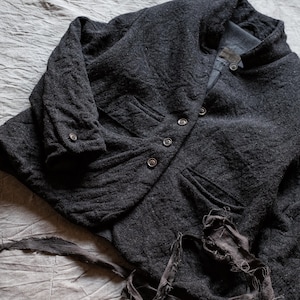 Könnte beinhalten: Eine dunkel anthrazitfarbene, texturierte Jacke mit Knopfleiste, Stehkragen und langen Ärmeln mit geknöpften Manschetten. Das Kleidungsstück verfügt über zwei Vordertaschen und ausgefranste, abgenutzte Stoffstreifen am unteren Saum.