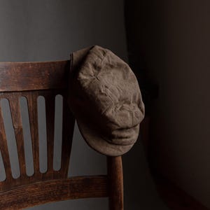 May include: A brown newsboy cap rests on the back of a wooden chair. The cap is made of a soft, textured material and has a rounded crown and a short brim. The chair is made of dark wood and has a slatted back.
