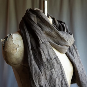 May include: A dark gray scarf draped over a mannequin. The scarf has a textured appearance with visible stitching and several square patches sewn onto it. The fabric appears to be lightweight, and the scarf is styled to showcase its drape.