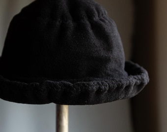 May include: A black cloche hat with a rounded crown and a small, textured brim. The hat is made of a soft, dark fabric and is displayed on a metal stand. The hat's design is simple and classic.