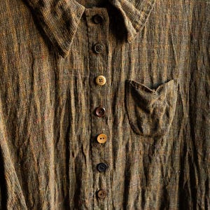 May include: A brown linen shirt with a button-up front and a small pocket on the left side. The shirt has a textured, woven fabric and features multiple brown buttons.