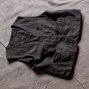 May include: A dark charcoal grey waistcoat featuring a V-neckline, a four-button front closure, and two lower welt pockets. The garment is constructed from a textured fabric and includes a small rectangular label stitched inside the neckline.