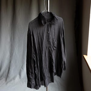 Pode incluir: Uma camisa cinza escuro com botões, gola e um bolso no peito, pendurada em um cabide de metal. A camisa de manga comprida tem uma aparência texturizada e ligeiramente desgastada, com linhas verticais visíveis no tecido.