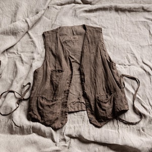 Könnte beinhalten: Eine braune Leinenweste mit zwei Vordertaschen und einem Knopfverschluss. Die Weste hat einen V-Ausschnitt und verstellbare Bänder an den Seiten. Das Kleidungsstück wird auf einem neutralfarbenen Stoffhintergrund präsentiert.