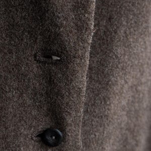May include: Close-up of a brown wool coat with a button and buttonhole detail. The coat has a textured surface and a dark button. The fabric appears to be a warm, neutral color.
