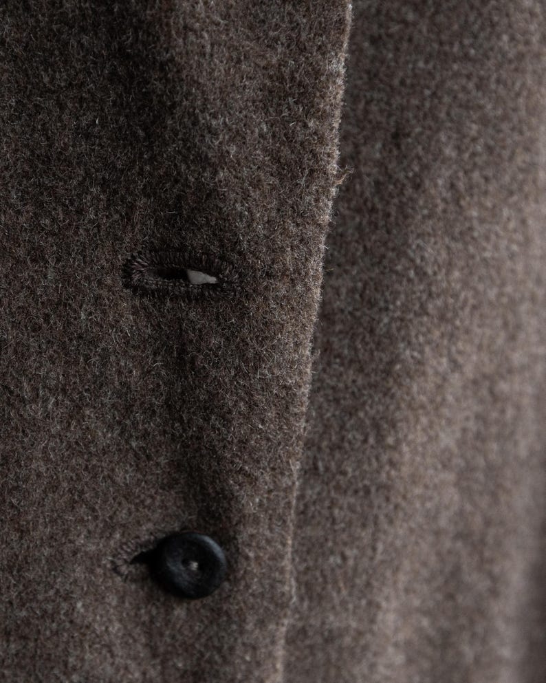 May include: Close-up of a brown wool coat with a button and buttonhole detail. The coat has a textured surface and a dark button. The fabric appears to be a warm, neutral color.