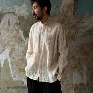 Puede incluir: Una camisa color crema de manga larga con cuello mandarín y cierre de botones. La camisa tiene un aspecto texturizado y ligeramente arrugado. La persona lleva pantalones negros. El fondo es una pared deteriorada con pintura descascarillada.
