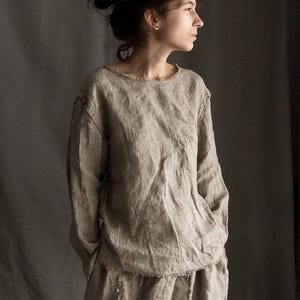 Könnte beinhalten: Eine Frau trägt ein locker sitzendes, hellbraunes Leinenoberteil mit einem rohen Ausschnitt und langen Ärmeln. Das Oberteil ist leicht überdimensioniert und hat eine lockere Passform. Die Frau trägt das Oberteil mit einer passenden Leinenhose.